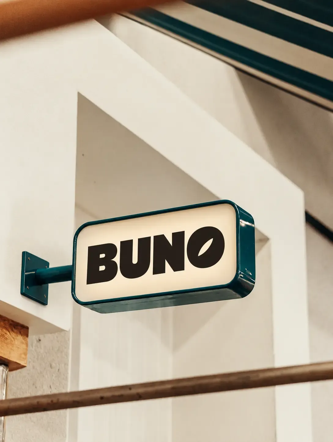 白い壁に置かれた青緑色のフレームのBuno店舗看板｜AI画像生成プロンプト